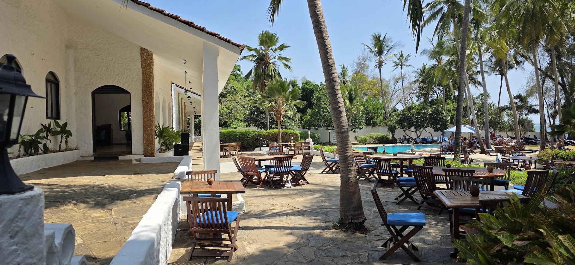 Diani Sea Lodge saulainā āra terase ar koka galdiem un zilām mīkstajām mēbelēm pie baseina un palmu dārza