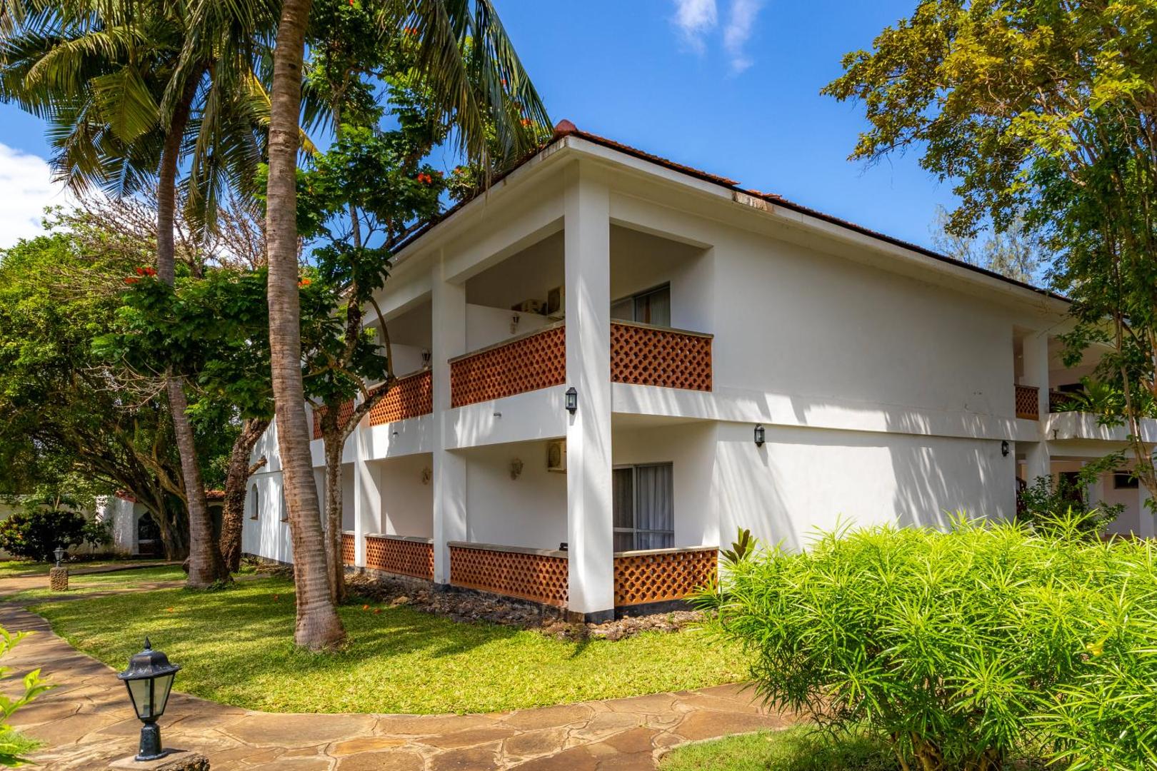 Diani Sea Lodge divstāvu viesnīcas korpuss ar balkoniem palmu dārzā — balta fasāde un akmens flīžu celiņš