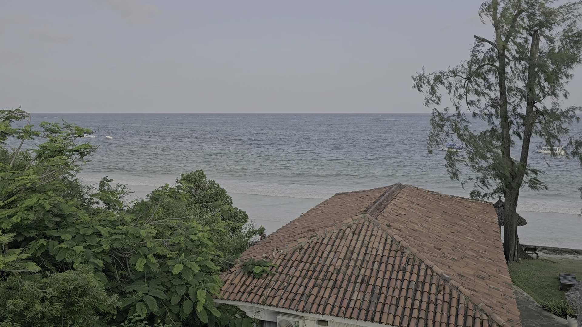 Skats no Diani Sea Lodge numura terases uz okeānu pāri sarkano dakstiņu jumtam un zaļojošiem kokiem Kenijas piekrastē