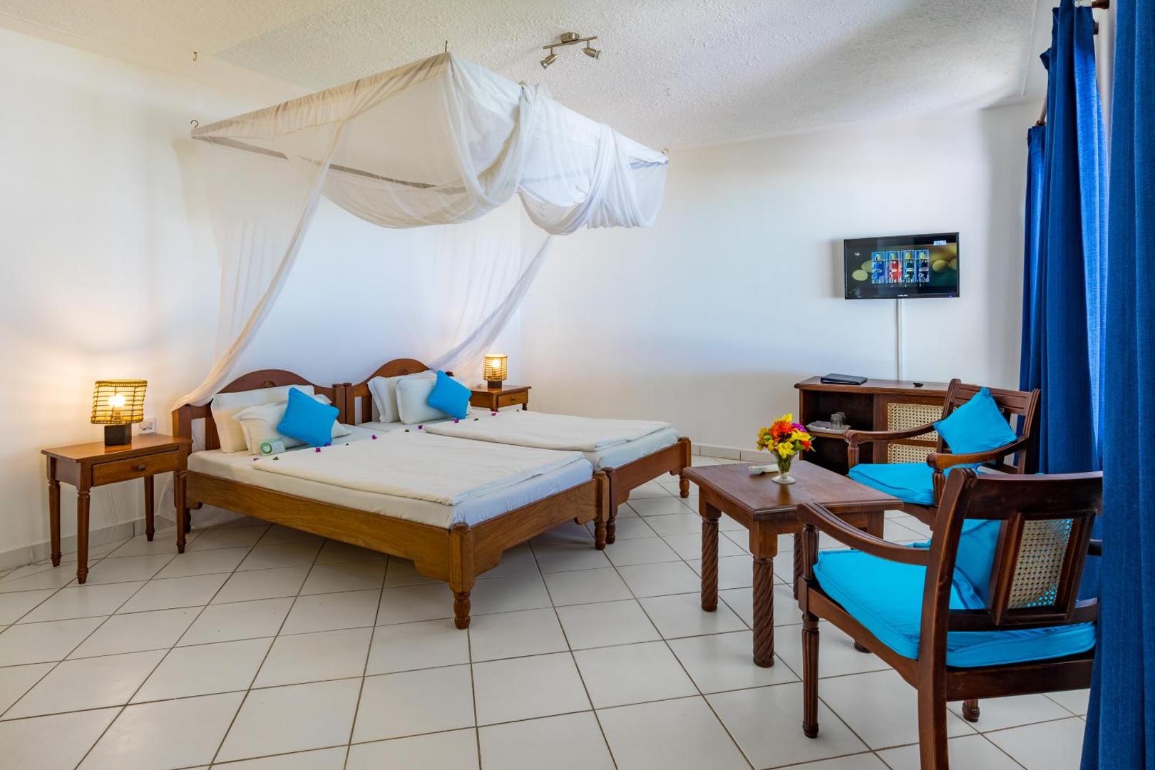 Diani Sea Lodge viesnīcas numurs ar divām gultām un moskītu tīklu Kenijā — gaišs interjers ar koka mēbelēm un ziliem akcentiem