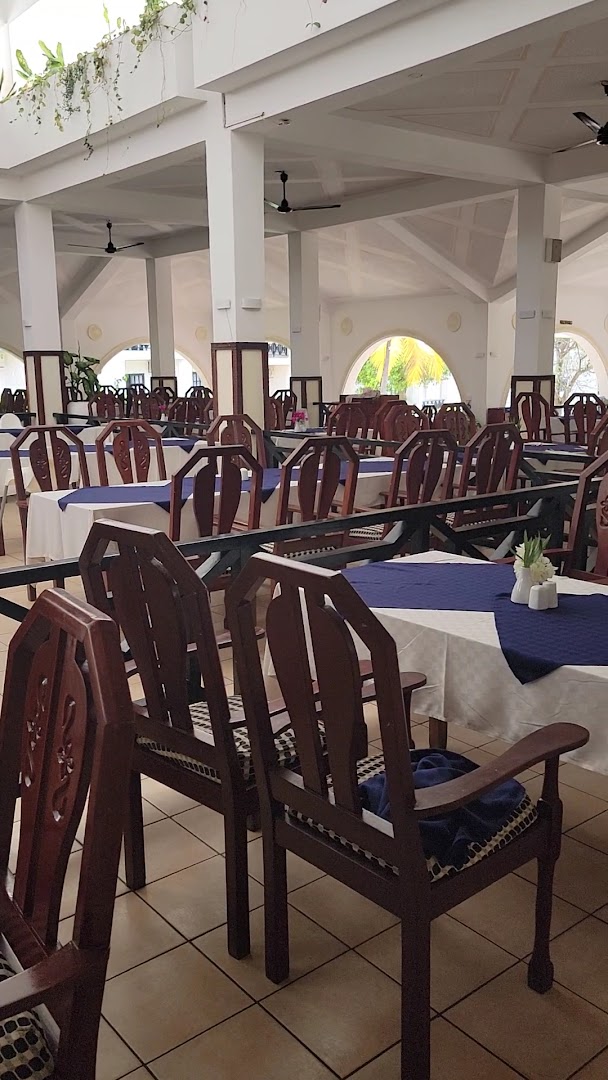 Diani Sea Lodge galvenā restorāna iekštelpas ar tumša koka krēsliem, baltiem galdautiem un augstiem griestiem