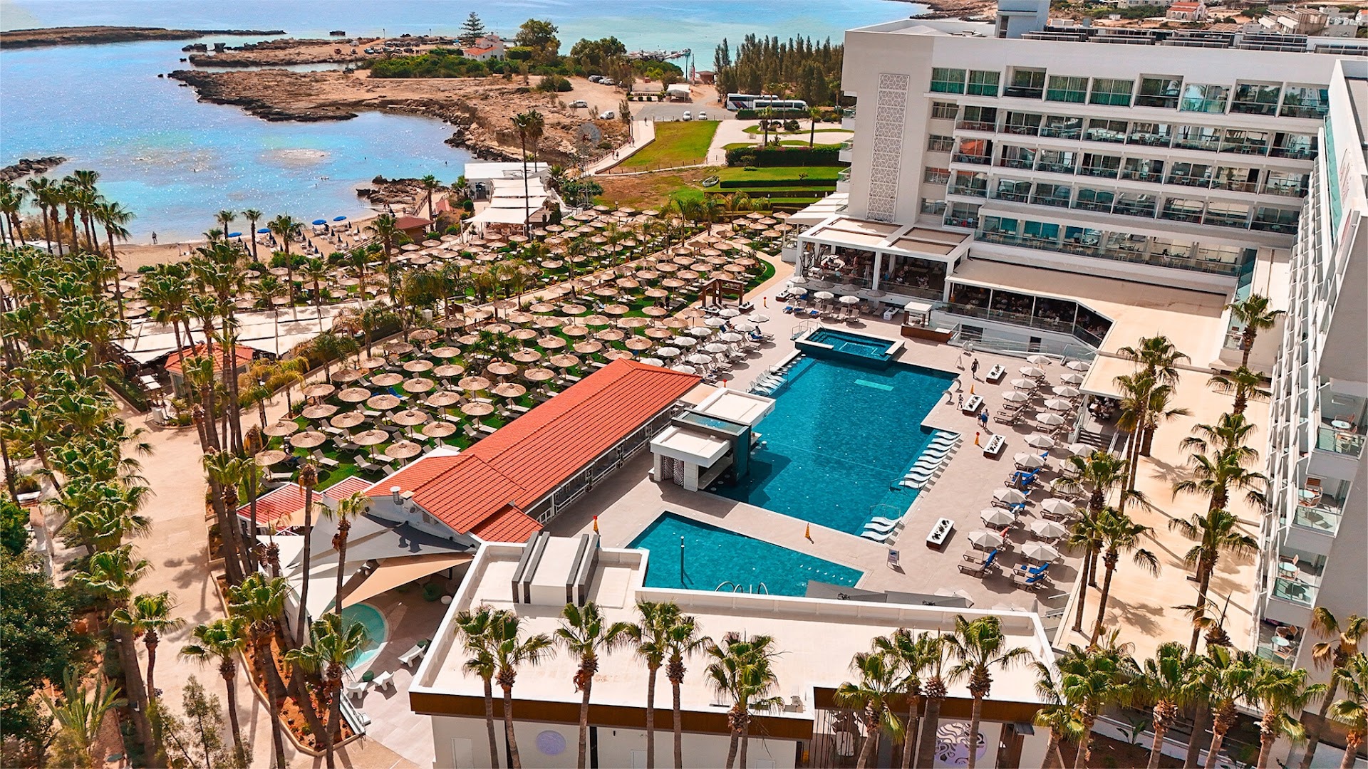 Cavo Maris Beach viesnīcas panorāmas skats Protarasā — moderna ēka, baseini un tieša piekļuve Vidusjūras līcim ar tirkīza ūdeni
