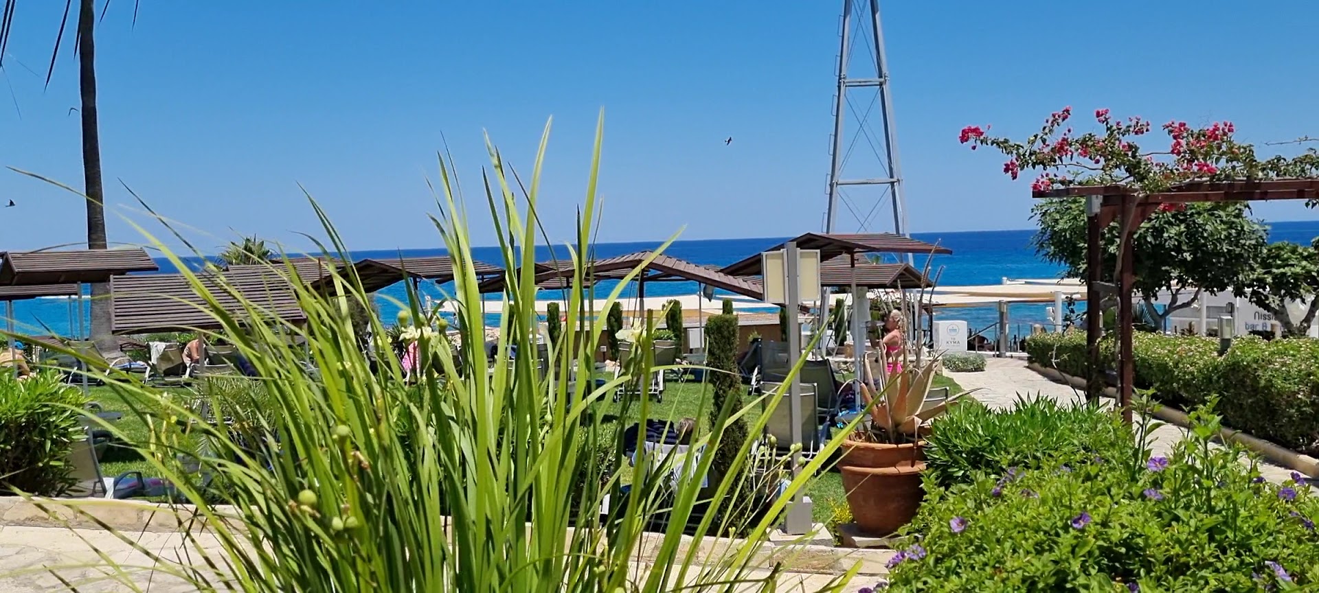 Skats uz Vidusjūru no Cavo Maris Beach viesnīcas dārza Protarasā — zaļš zāliens ar saulessargiem, ziedoši augi un tirkīzzils ūdens