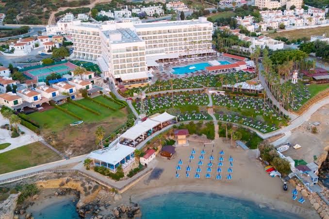 Cavo Maris Beach Hotel aerofoto — viesnīcas ēka, tenisa korti, baseins un tieša piekļuve smilšu pludmalei Protarasā