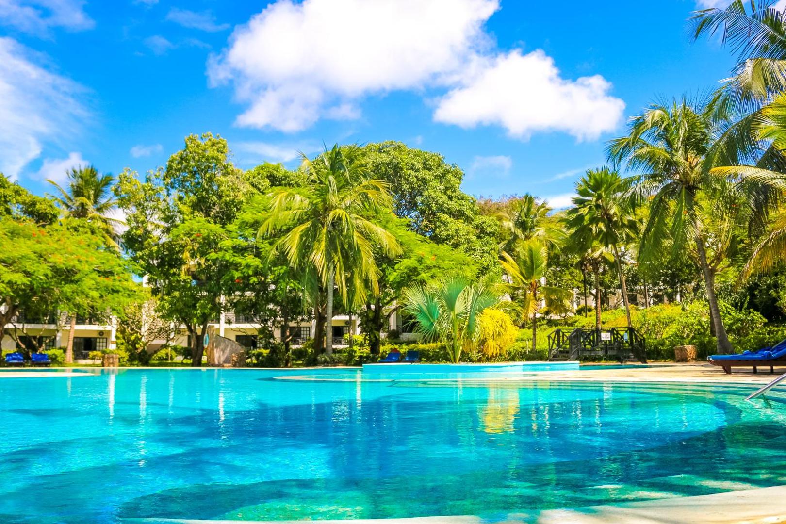 Diani Sea Resort dzidrais baseina ūdens tuvplānā ar palmu atspulgiem, dārza apstādījumiem un koši zilām debesīm fonā.