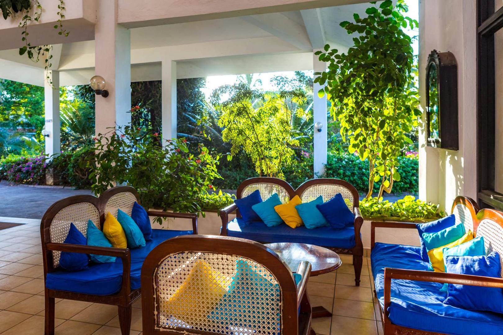 Diani Sea Resort atpūtas zona ar ziliem dīvāniem un dzelteniem spilveniem atvērtā tipa terasē, ko ieskauj zaļi augi