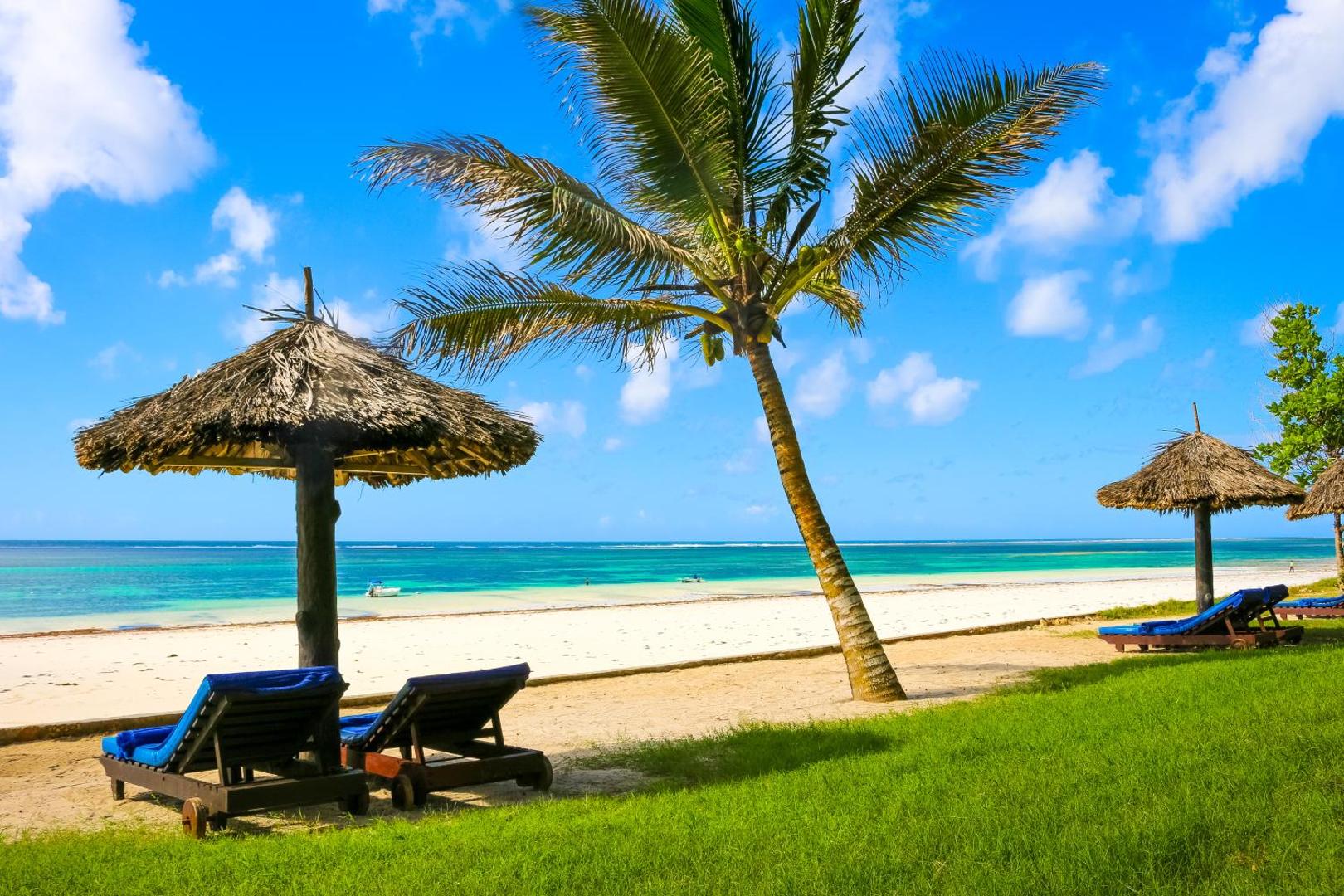 Atpūtas krēsli zem salmu saulessargiem zālienā pie Diani Sea Resort pludmales — skats uz okeānu un palmu