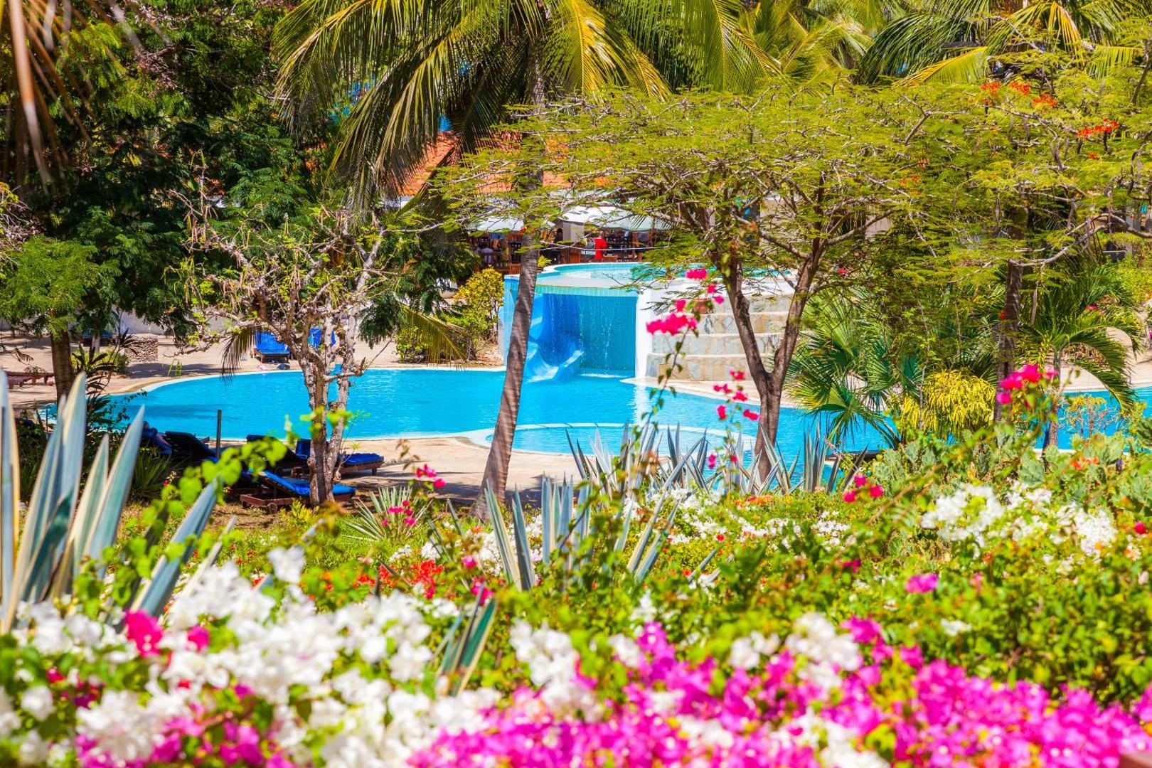 Diani Sea Resort ziedošs dārzs ar košiem ziediem priekšplānā, skatu uz baseinu, slidkalniņu un blīvu tropu veģetāciju.