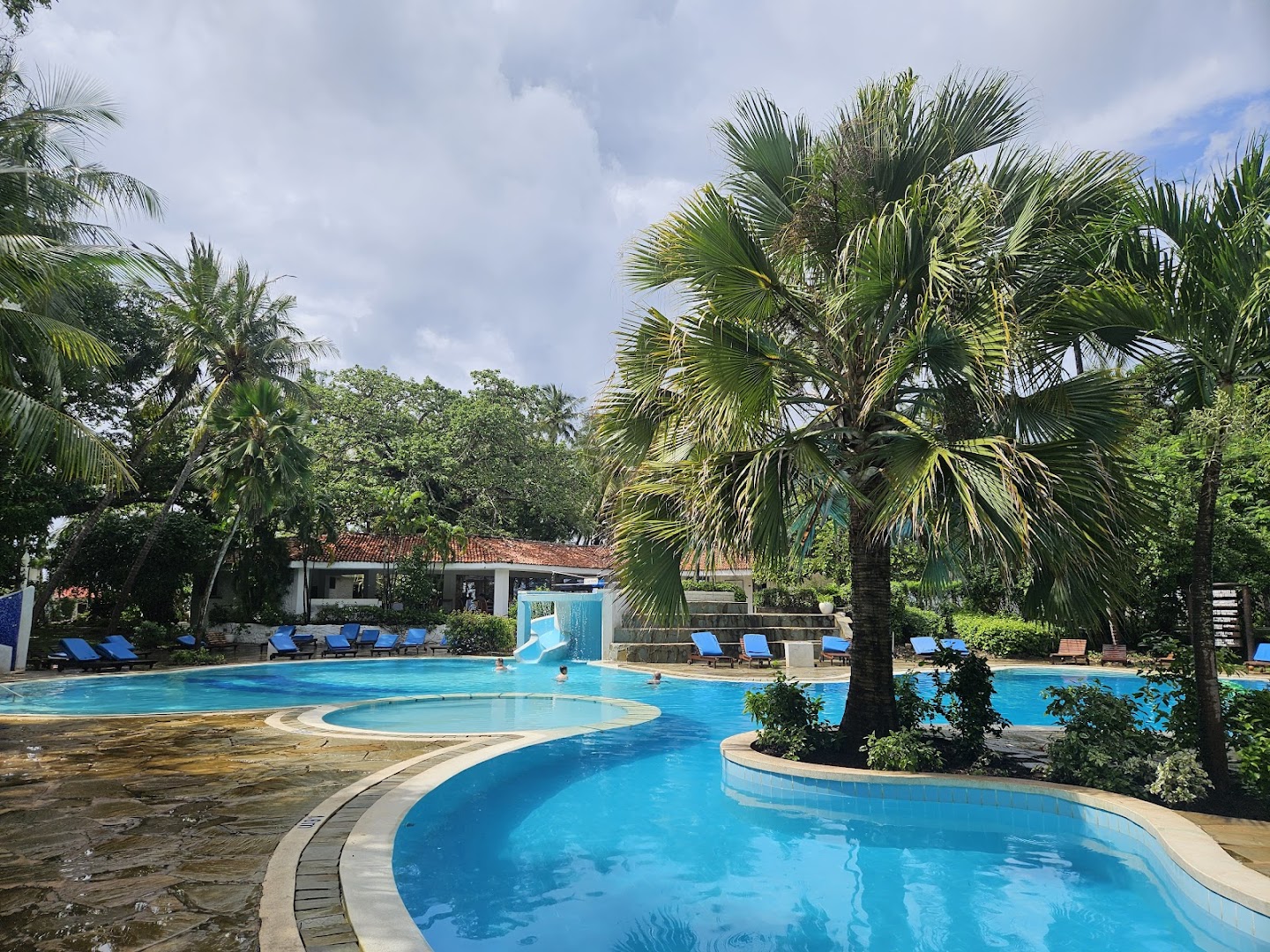 Diani Sea Resort āra baseins ar nelielu slidkalniņu, ziliem sauļošanās krēsliem un eksotiskām palmām mākoņainā dienas laikā.