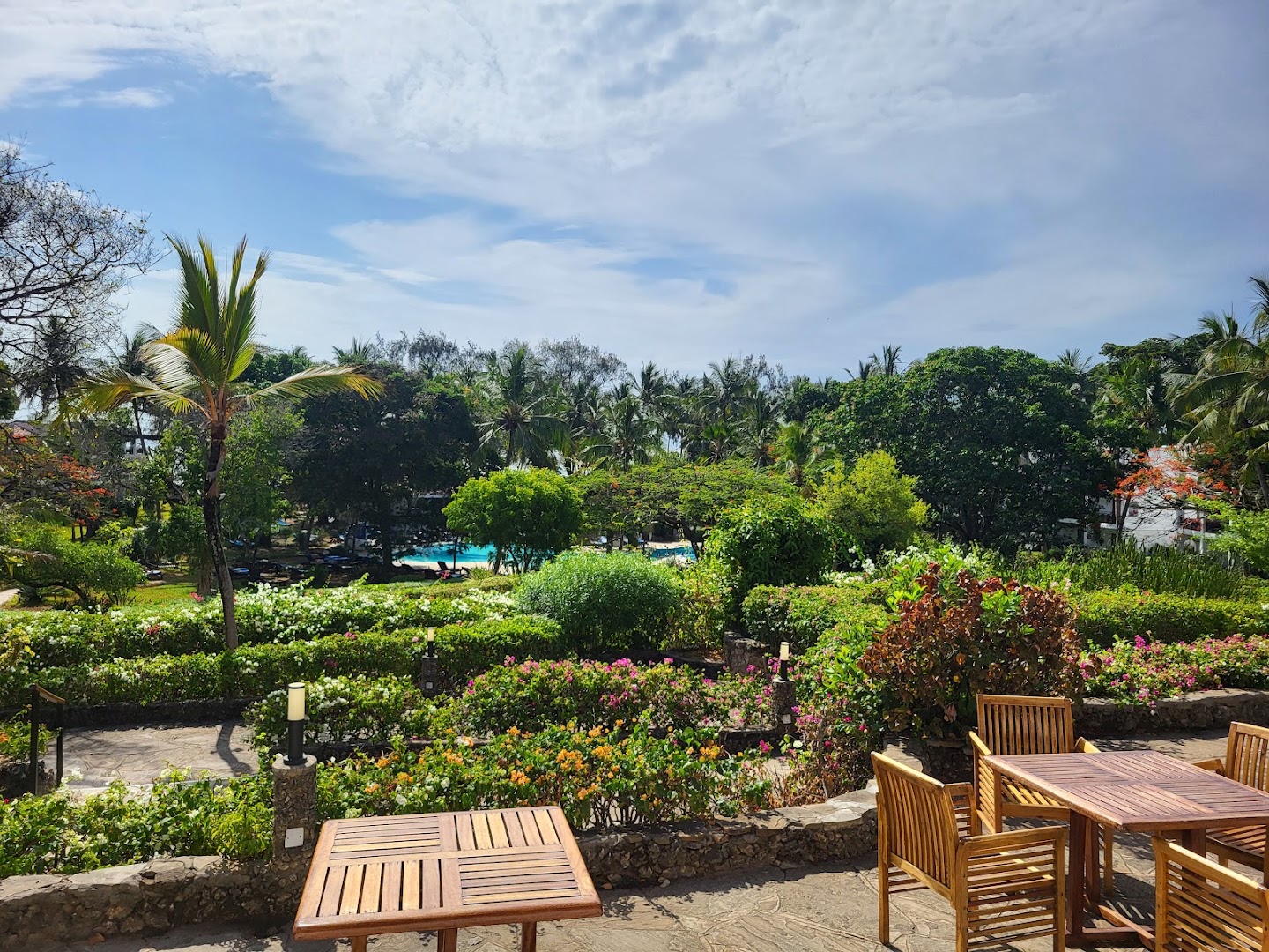 Diani Sea Resort āra terase ar koka galdiem un krēsliem, ko ieskauj ziedoši krūmi un tropu dārzs ar skatu uz baseinu