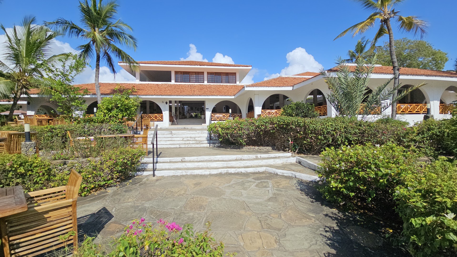 Diani Sea Resort galvenā ēka ar terasi un koka mēbelēm Kenijā — balta arhitektūra ar sarkaniem dakstiņu jumtiem un tropu augiem