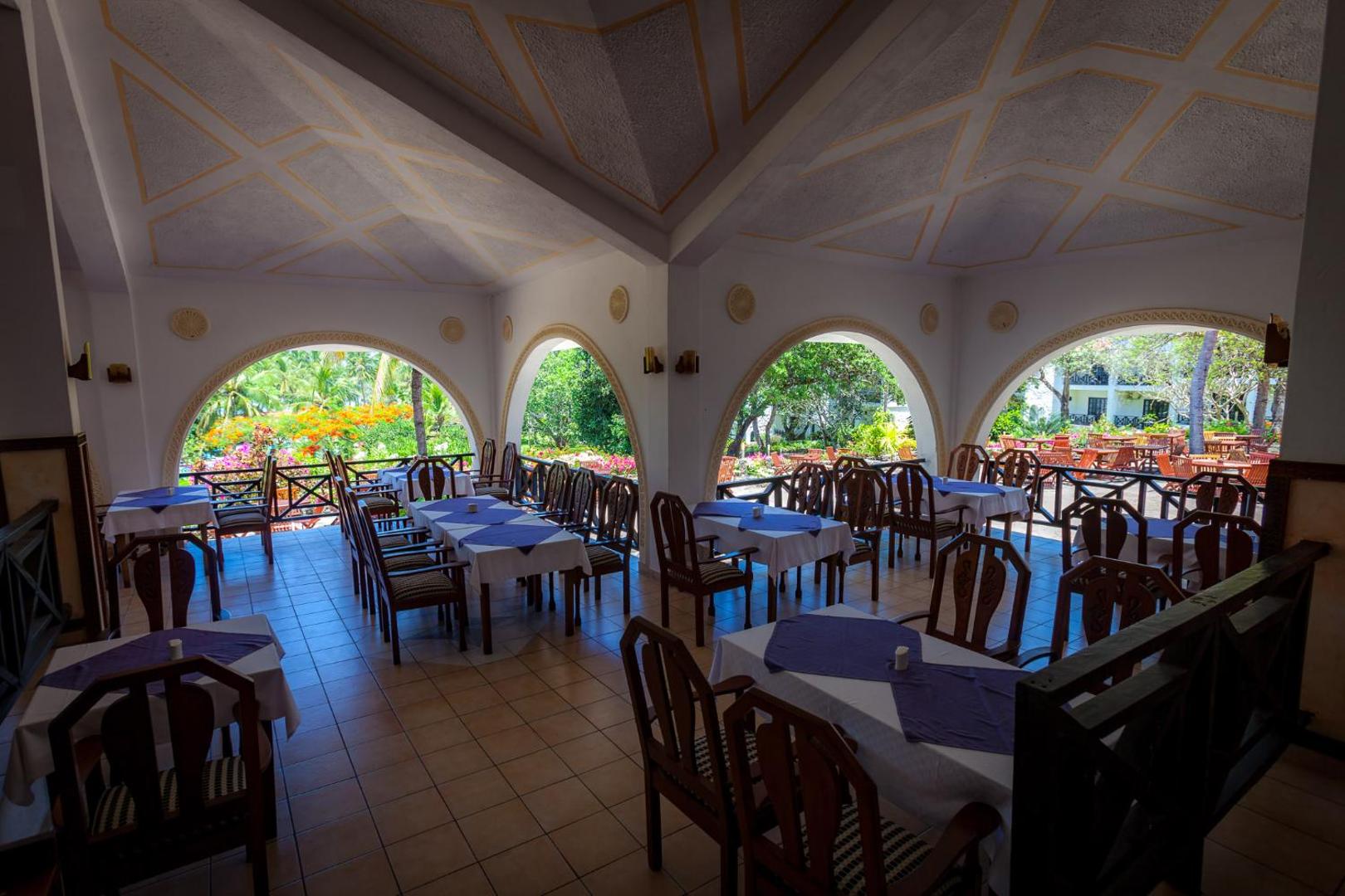 Diani Sea Resort restorāna iekštelpas ar arkveida logiem, klātiem galdiem un skatu uz viesnīcas zaļo teritoriju un dārzu