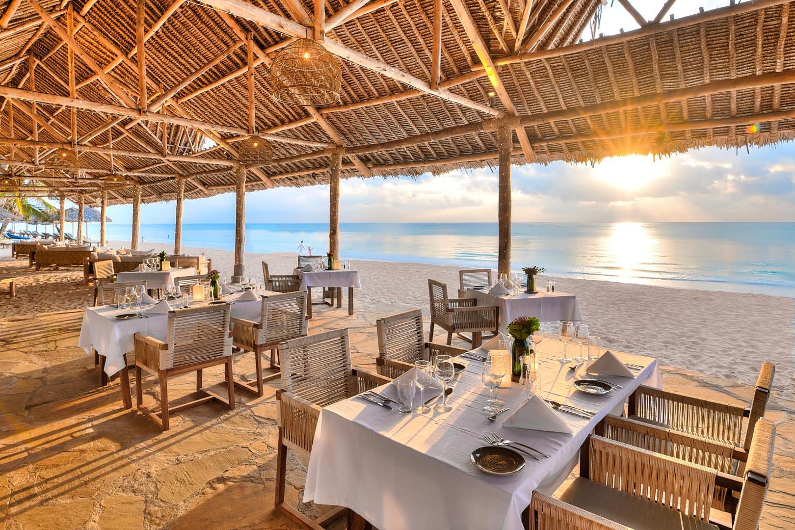Diamond Leisure Beach & Golf Resort restorāns pludmalē saulrieta laikā ar klātiem galdiem zem tradicionāla niedru jumta Zanzibārā