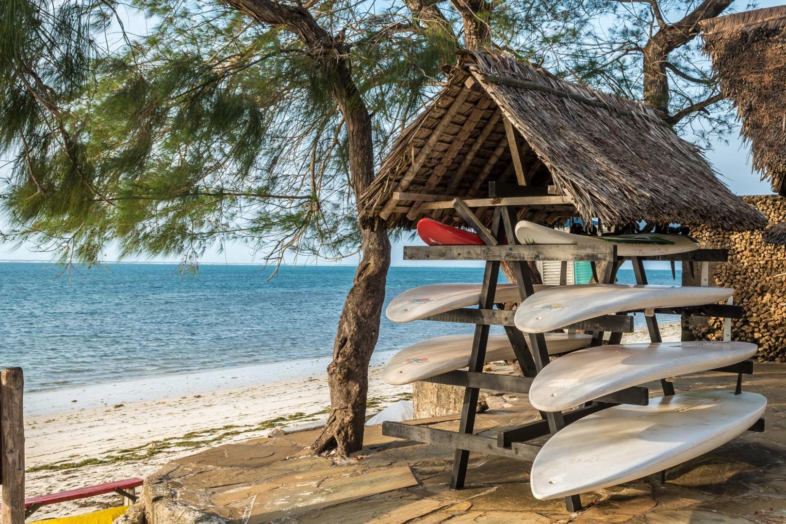Serena Beach Resort & Spa sērfošanas dēļu novietne zem salmu jumta okeāna piekrastē ar baltu smilšu pludmali fonā.