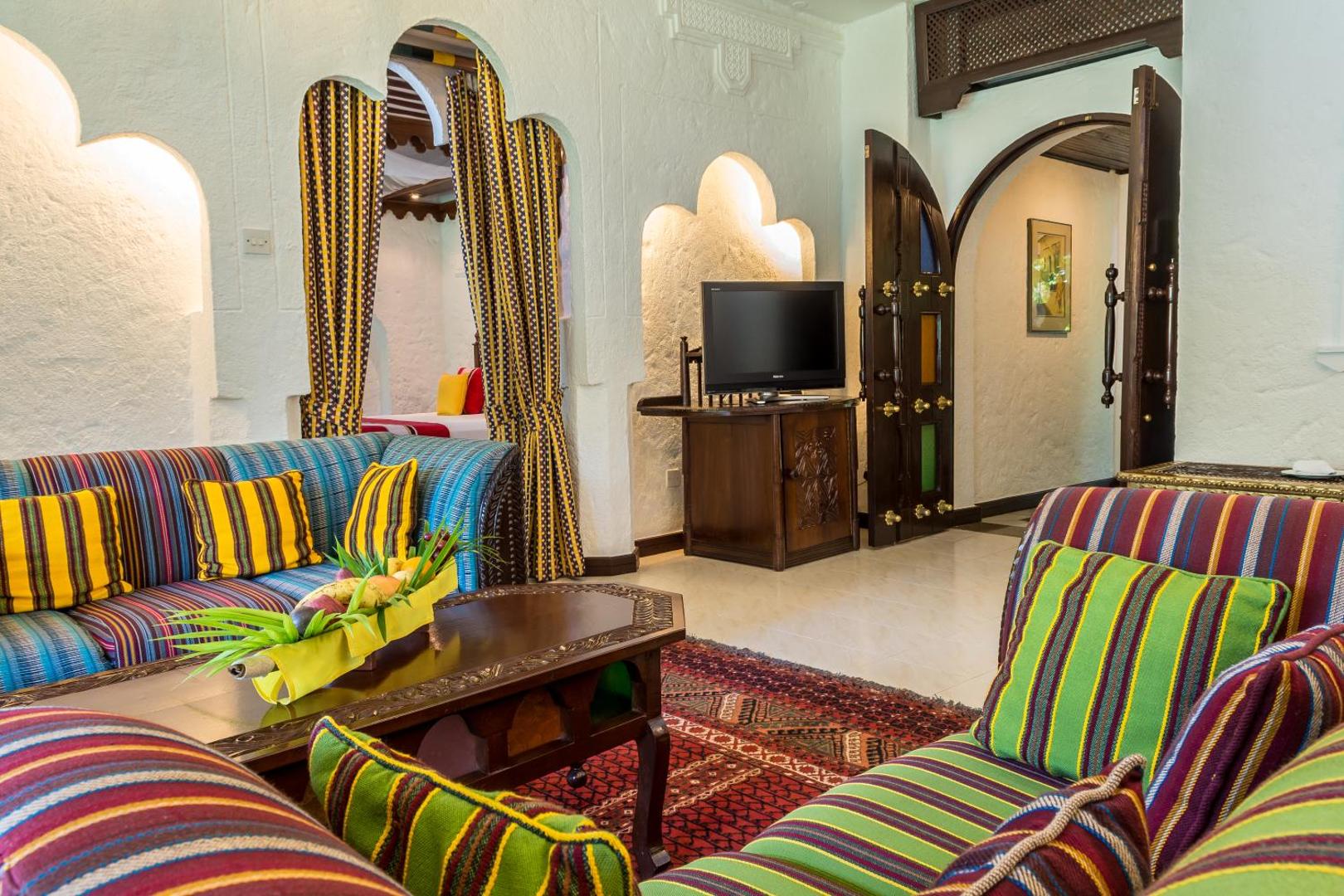 Serena Beach Resort & Spa viesnīcas numura viesistabas zona ar krāsainiem dīvāniem, koka galdiņu un tradicionālām arkām Mombasā