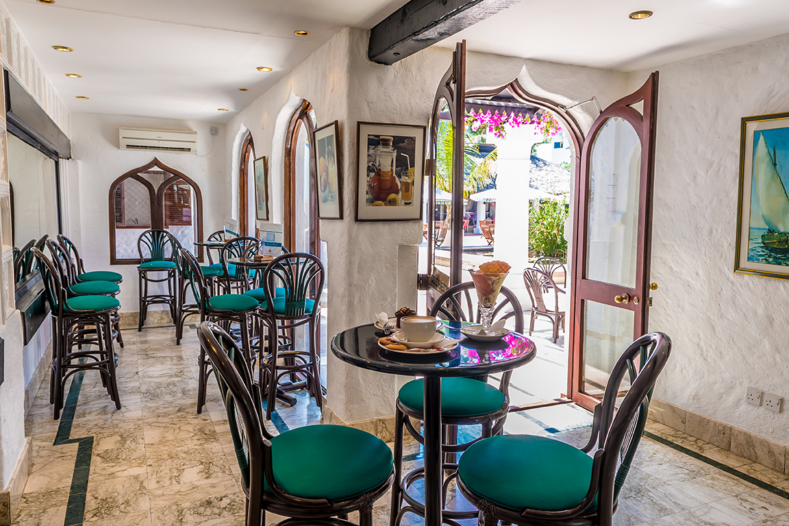 Gaiša kafejnīca Serena Beach Resort & Spa viesnīcā ar marmora grīdu, tirkīza krēsliem un skatu uz dārzu caur arkveida durvīm.