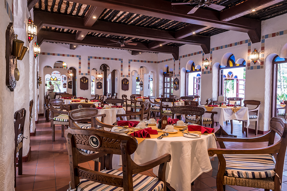Serena Beach Resort & Spa restorāna interjers ar tradicionālu koka siju griestiem, arkveida logiem un klātiem galdiem Mombasā
