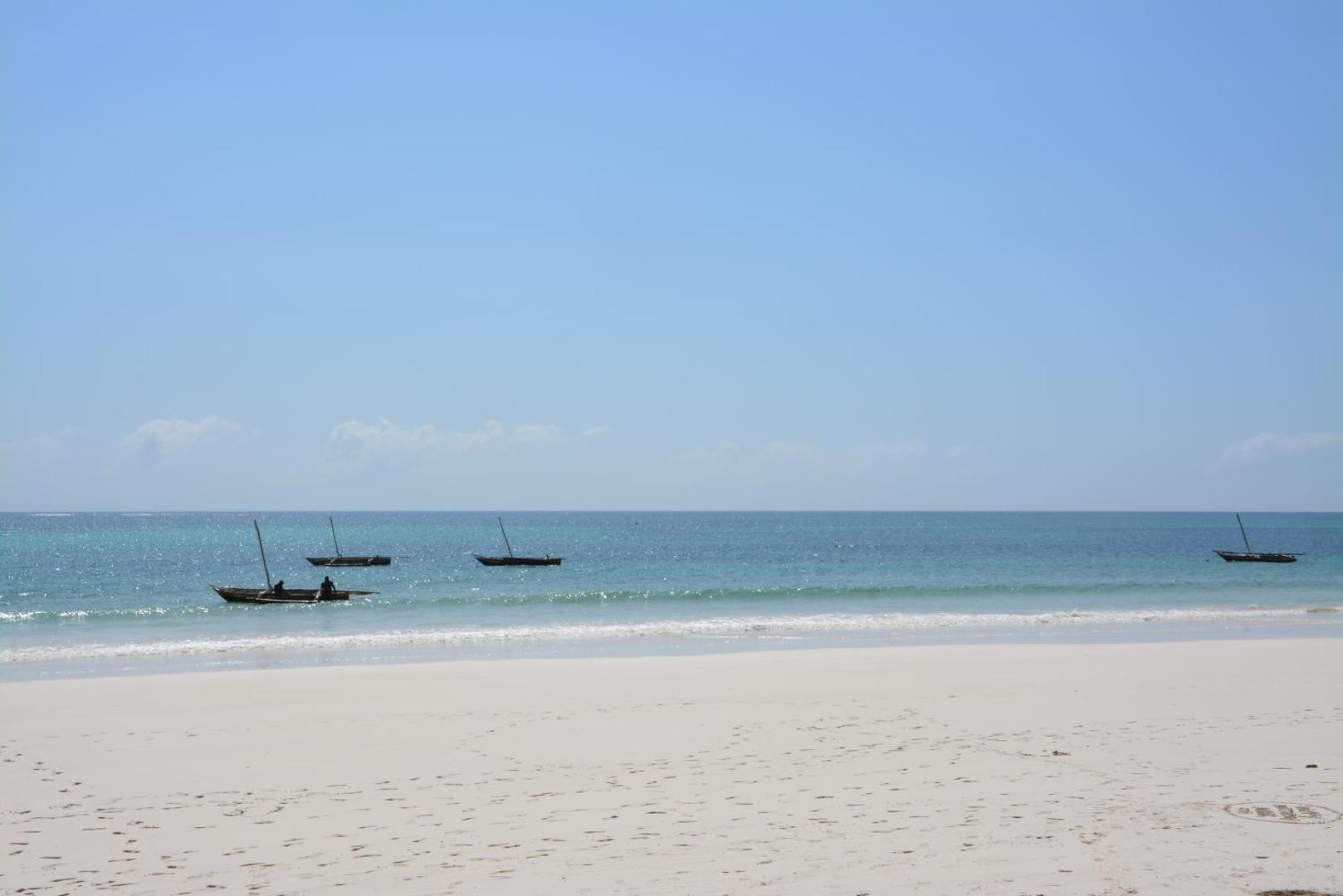 Neptune Palm Beach Boutique Resort & Spa baltu smilšu pludmale Zanzibārā ar tradicionālām koka laivām tirkīzzilā Indijas okeāna ūdenī.