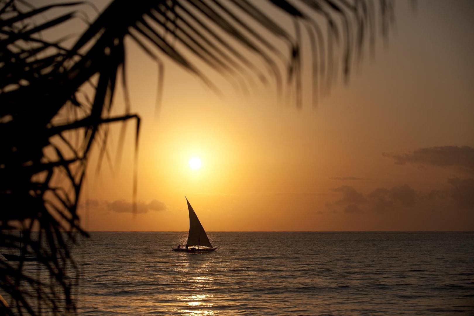 Neptune Palm Beach Boutique Resort & Spa skats uz saulrietu okeānā ar tradicionālo buru laivu un palmu siluetiem piekrastē