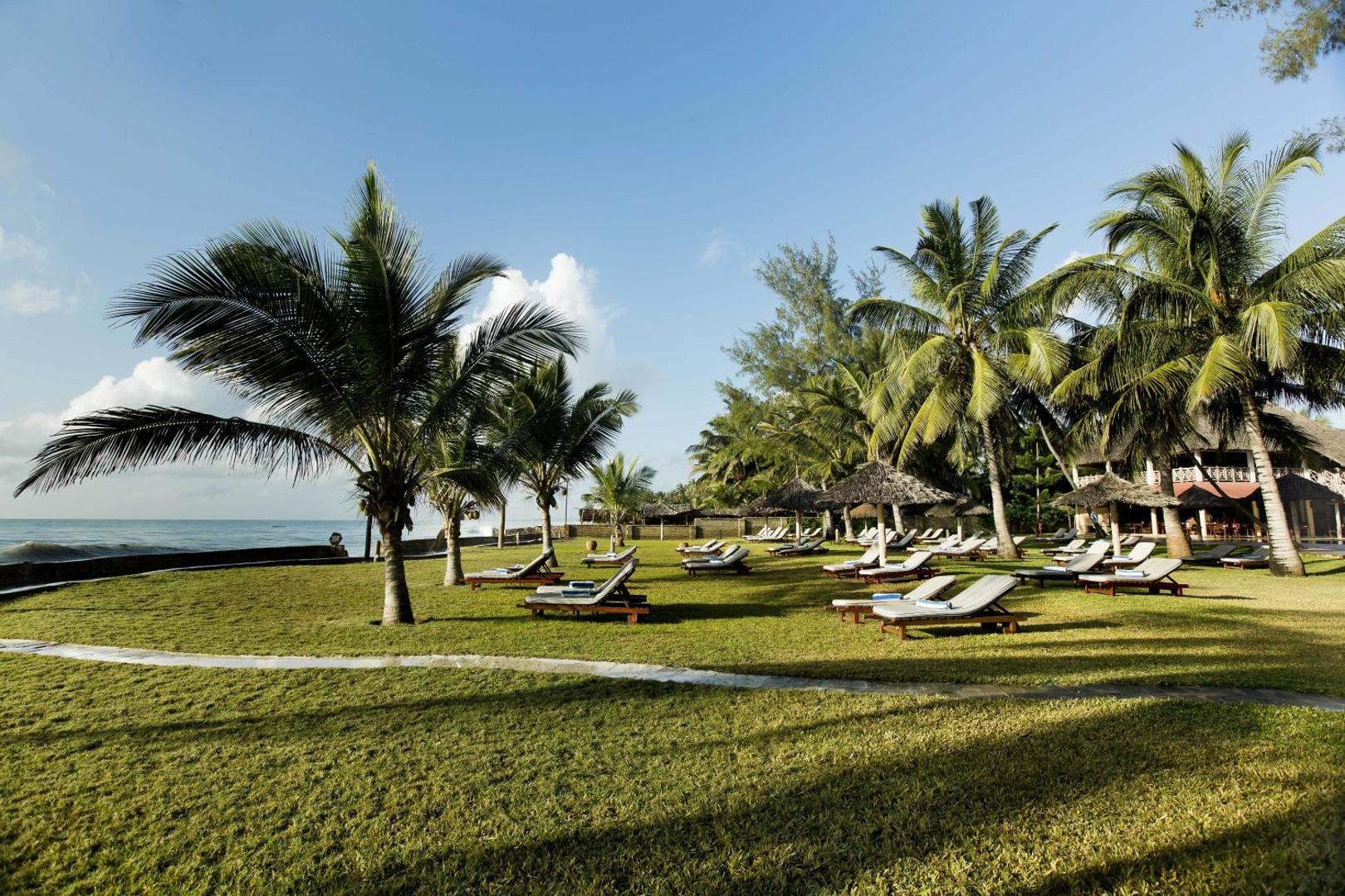 Neptune Palm Beach Boutique Resort & Spa dārzs ar zālāju, palmām un sauļošanās krēsliem okeāna piekrastē Zanzibārā, Tanzānijā.