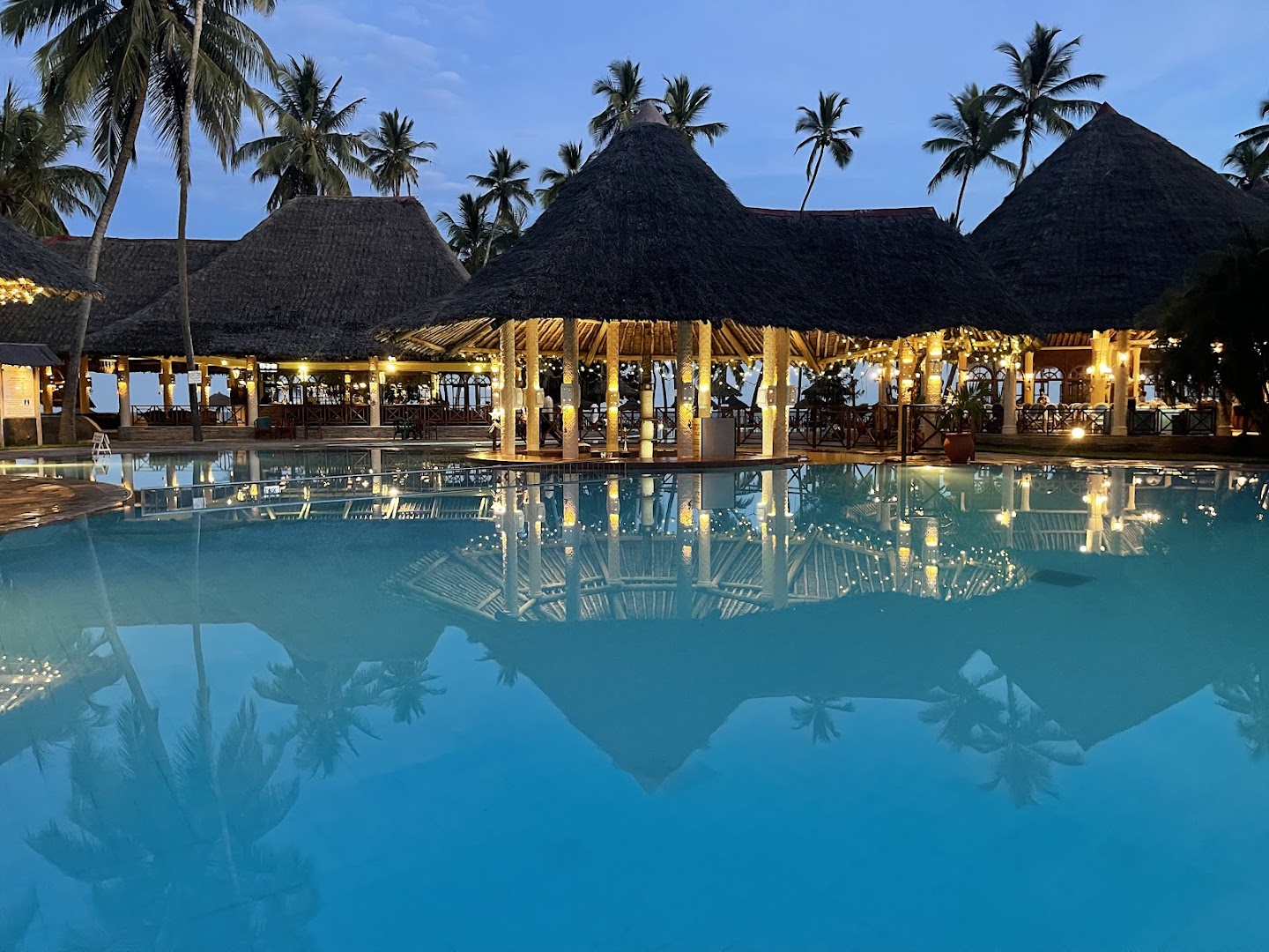 Neptune Palm Beach Boutique Resort & Spa baseina bārs krēslas stundā ar atspulgu ūdenī un palmām fonā, Kenija