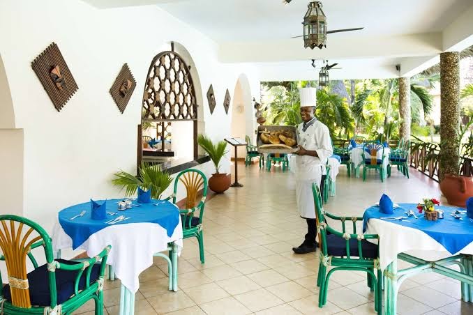 Neptune Palm Beach Boutique Resort & Spa restorāna terase ar balti klātiem galdiem un viesmīli, kurš pasniedz svaigu maizi