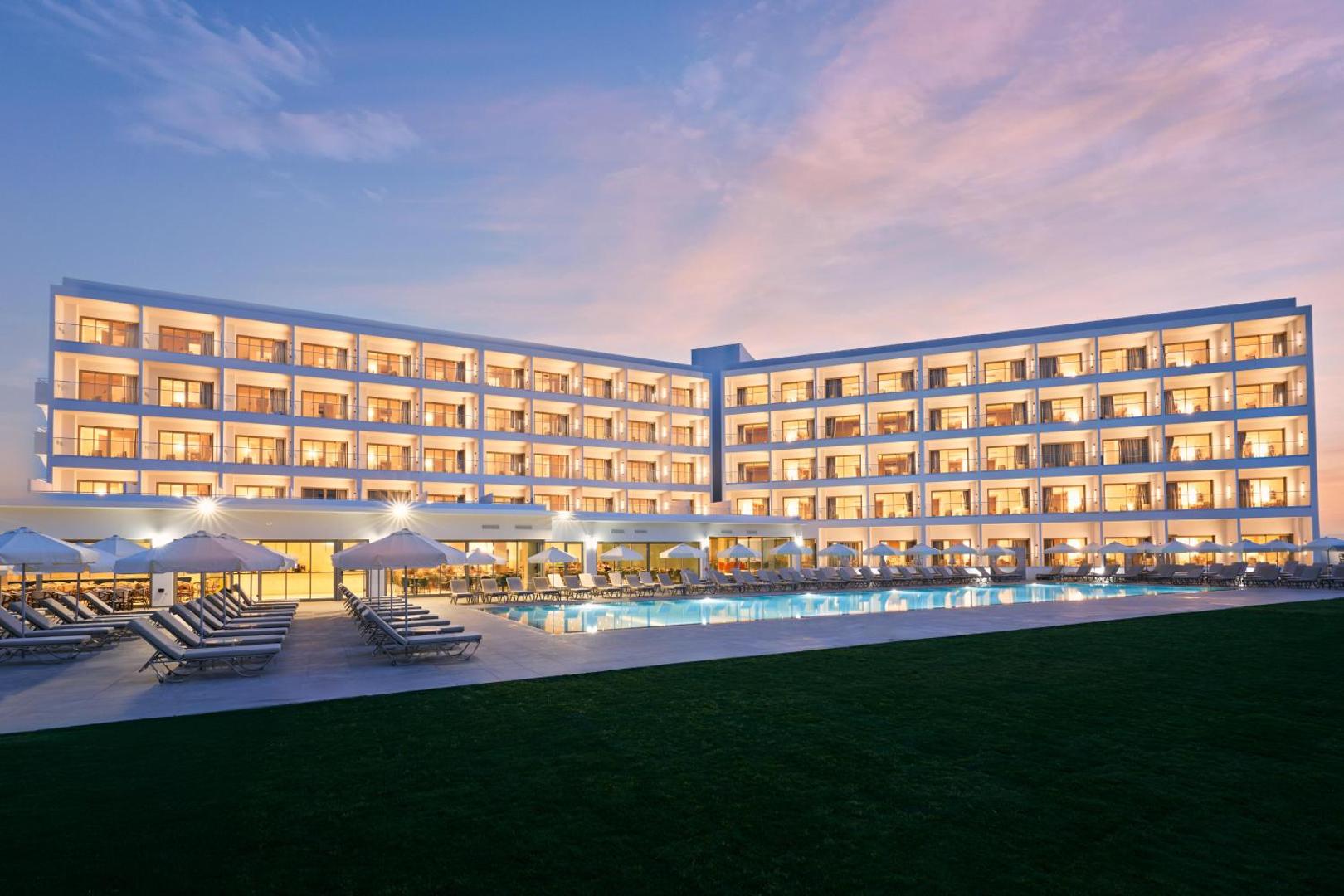 Maris Grand Waterpark Resort galvenā ēka un āra baseins saulrietā Rodas salā — apgaismota fasāde, sauļošanās krēsli un zāliens