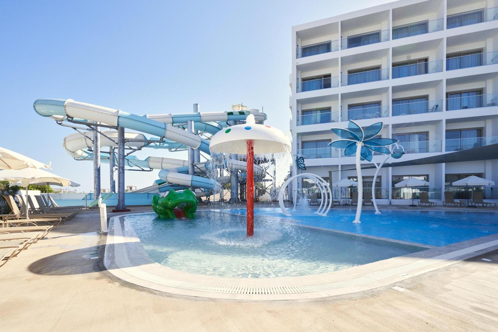 Maris Grand Waterpark Resort bērnu baseins ar strūklakām, sēni un vardi, fonā viesnīcas ēka un lielie slidkalniņi Kiprā