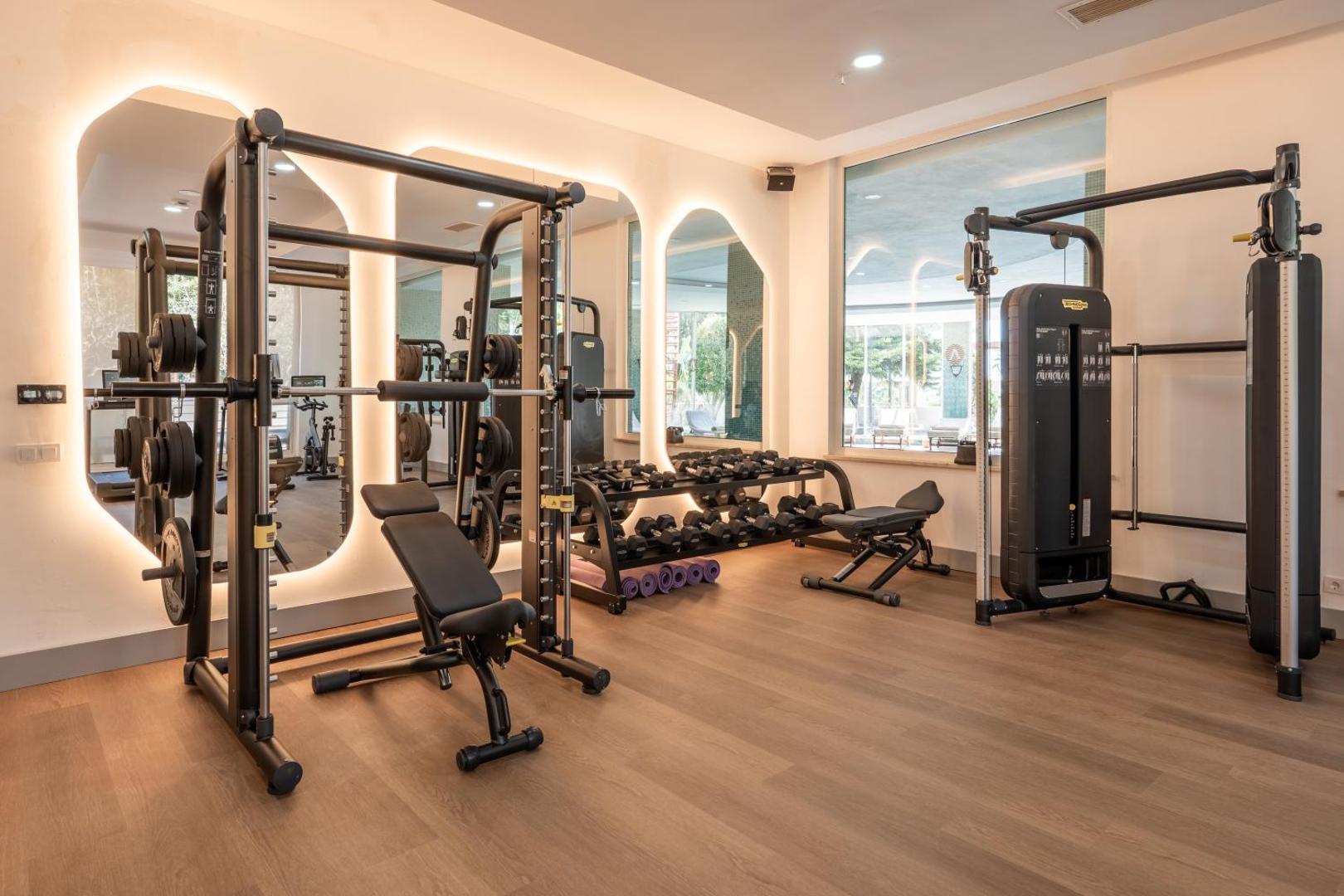Adalya Bliss moderns fitness centrs ar trenažieriem, svariem un lieliem spoguļiem gaišā telpā