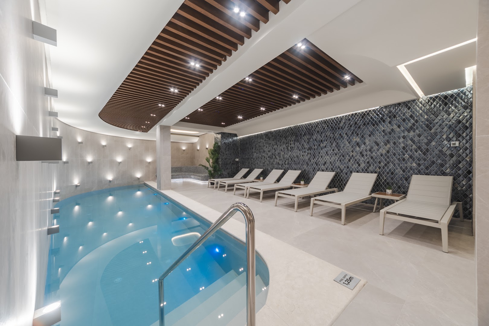 Viesnīcas Katamare iekštelpu spa baseins ar zilu apgaismojumu, atpūtas krēsliem un modernu griestu apdari relaksācijai.