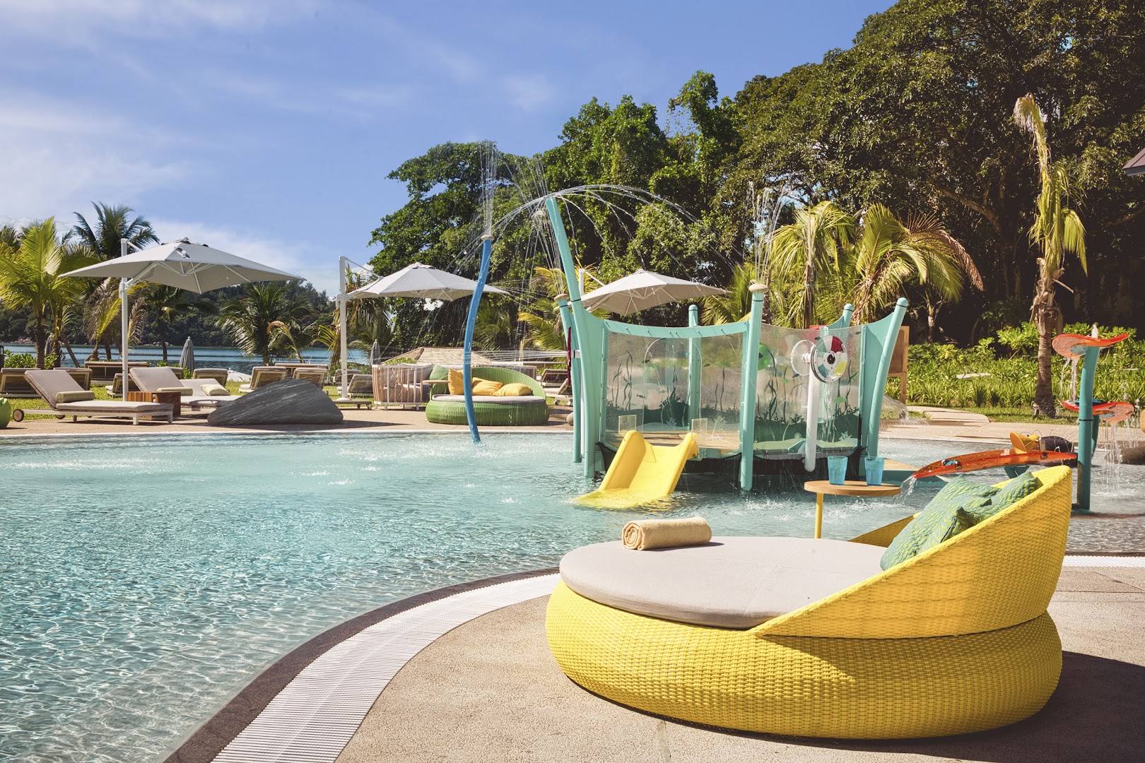 Club Med Seychelles bērnu baseins ar dzeltenu slidkalniņu, ūdens strūklakām un ērtiem sauļošanās krēsliem ģimenes atpūtai