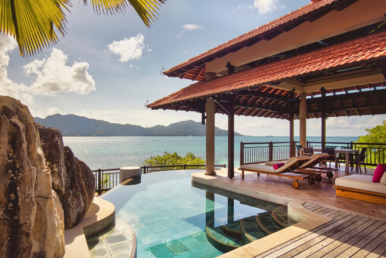 Club Med Seychelles bezgalības baseins piekrastē ar skatu uz okeānu un kalniem, sauļošanās krēsli zem nojumes