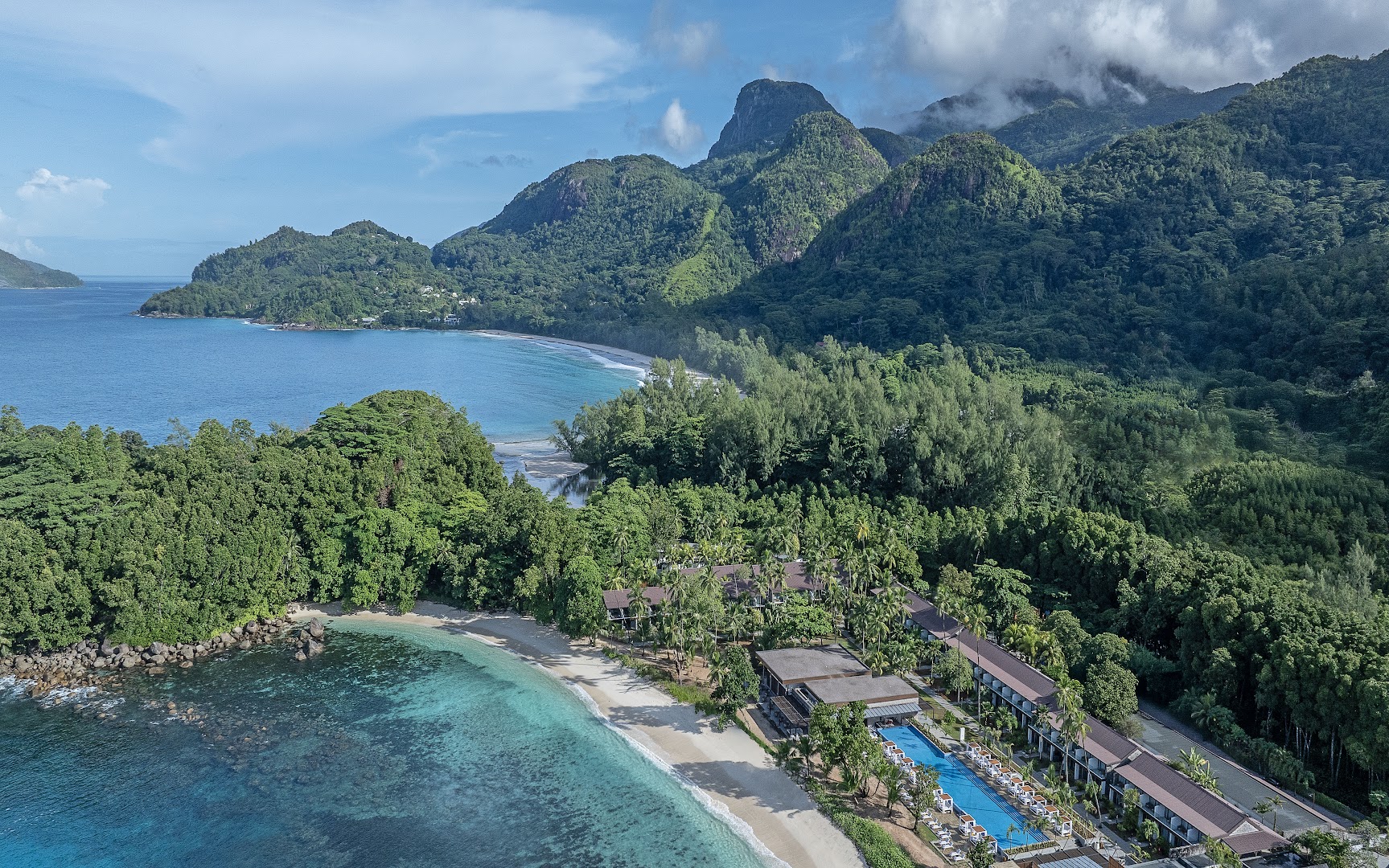 Panorāmas skats uz Avani+ Barbarons Seychelles piekrasti, kur viesnīcas ēkas izvietotas gar smilšu pludmali un garu āra baseinu
