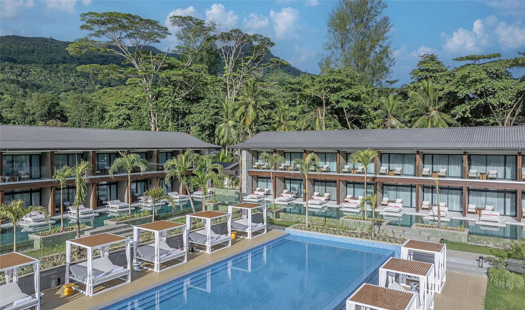 Avani+ Barbarons Seychelles Resort kopskats uz āra baseinu un divstāvu korpusiem ar numuriem, ko ieskauj palmas un kalni Seišelu salās
