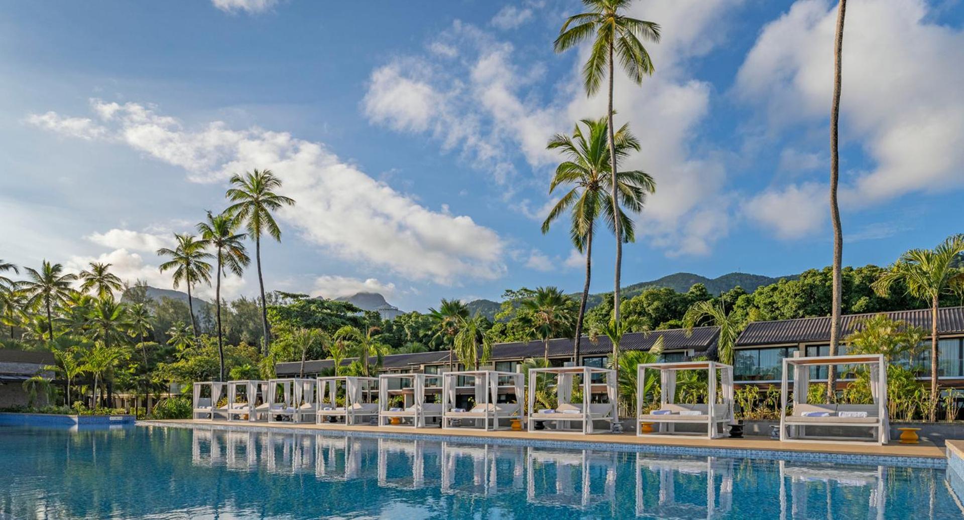 Avani+ Barbarons Seychelles āra baseins ar stilīgām atpūtas nojumēm un augstām palmām pret skaidrām debesīm Seišelu salu kūrortā