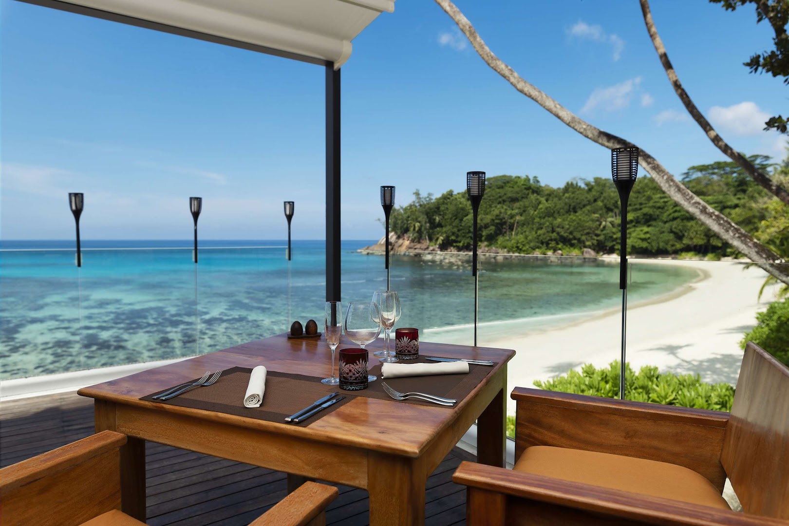 Avani+ Barbarons Seychelles restorāna terase ar klātu koka galdu diviem, skatu uz balto smilšu pludmali un tirkīza ūdeni saulainā dienā.