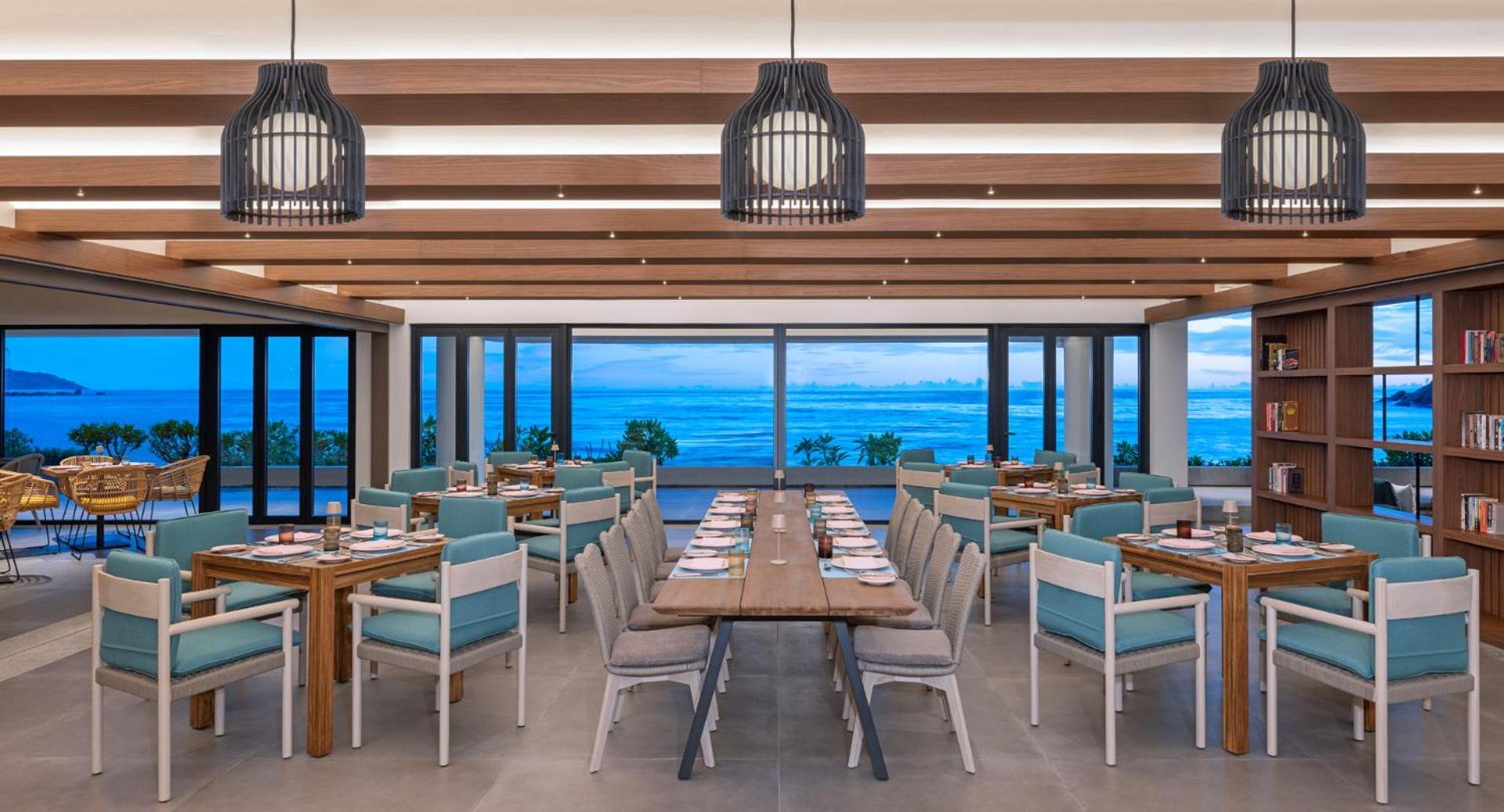 Avani+ Barbarons Seychelles moderns viesnīcas restorāns ar koka griestu sijām, stilīgiem gaismekļiem un panorāmas skatu uz okeānu krēslas stundā.