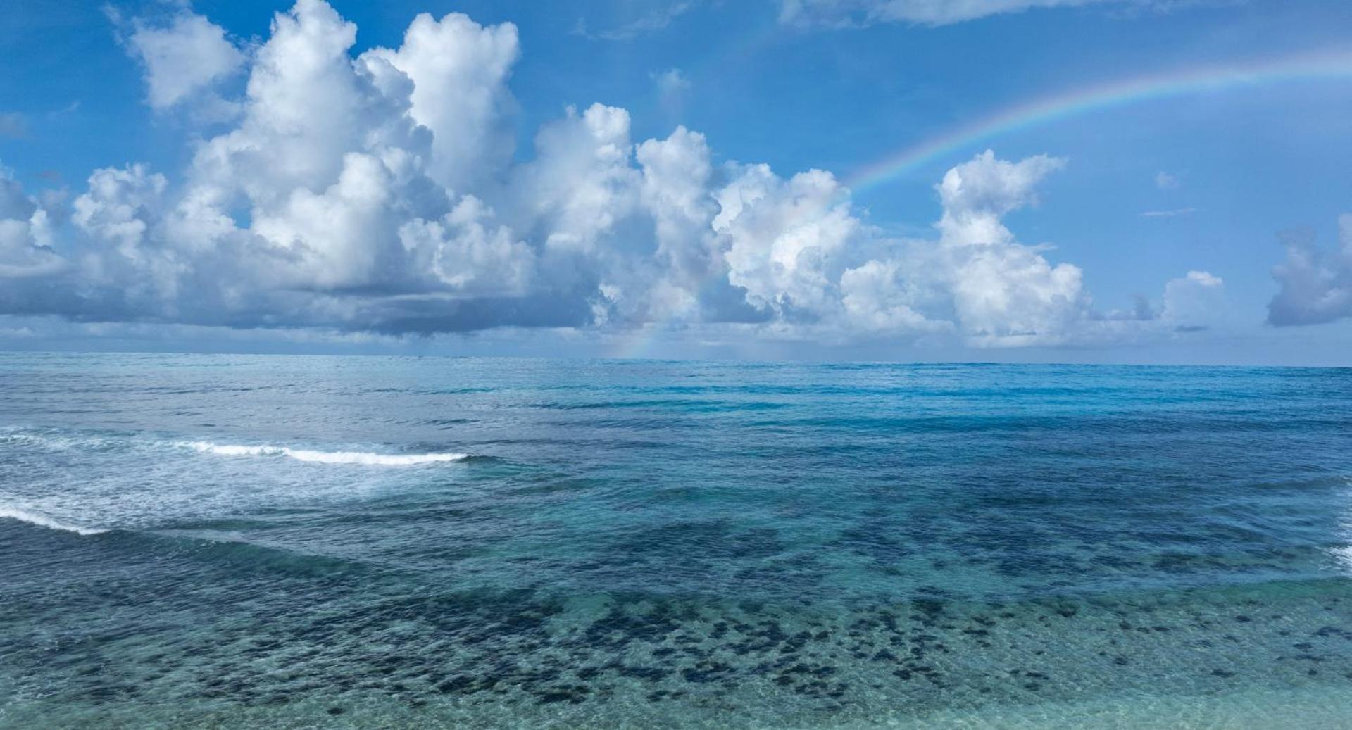 Avani+ Barbarons Seychelles skats uz Indijas okeāna viļņiem zem zilām debesīm ar gubumākoņiem un krāsainu varavīksni pie horizonta.