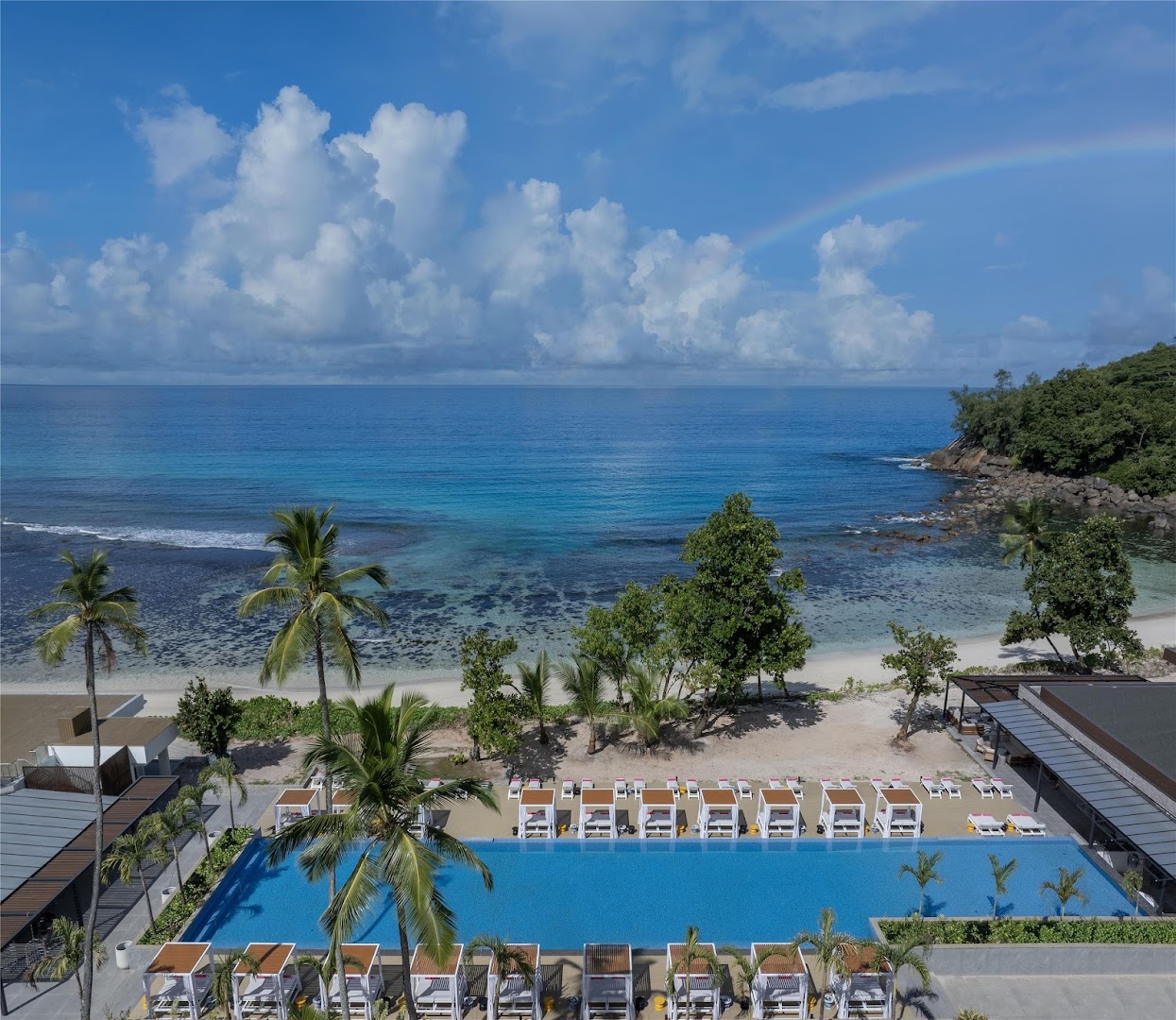 Avani+ Barbarons Seychelles panorāmas skats no augšas uz zilo okeānu, pludmali un baseinu ar varavīksni debesīs Seišelu salu piekrastē