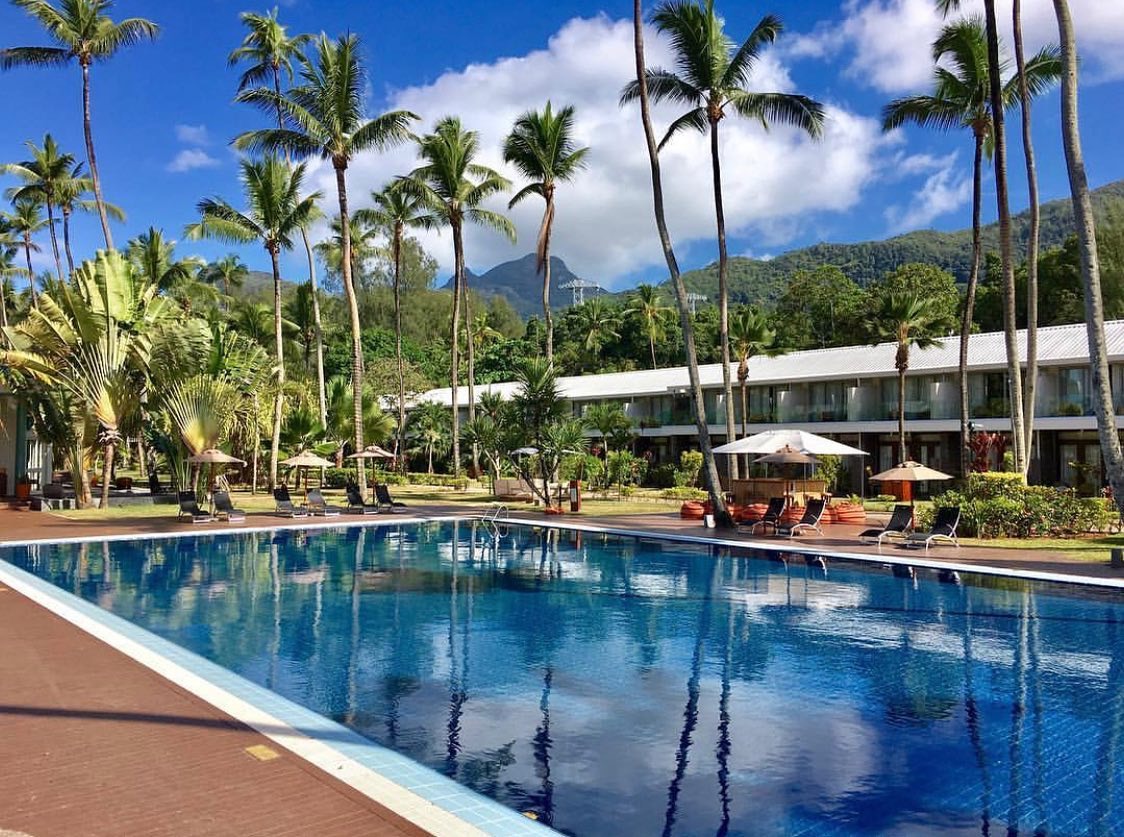 Avani+ Barbarons Seychelles galvenais āra baseins ar palmām un kalnu fonā saulainā dienā, atpūtas krēsli un saulessargi piekrastē