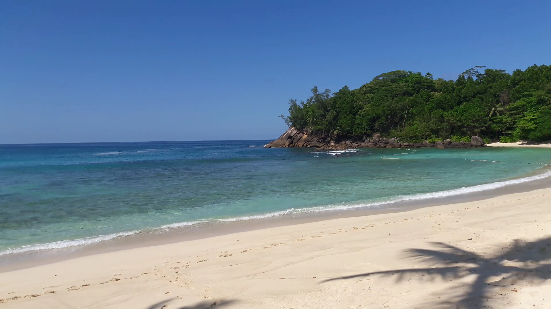 Gleznains skats uz Avani+ Barbarons Seychelles pludmales līci — baltas smiltis, dzidrs ūdens un zaļojošs kalns fonā piekrastē