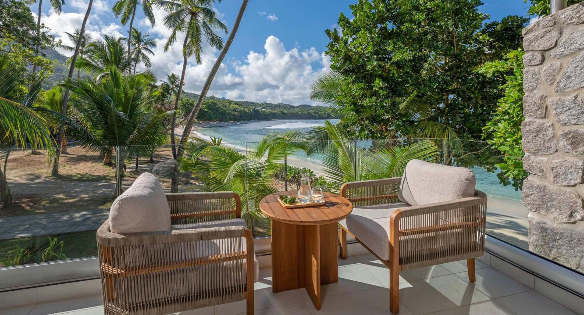 Avani+ Barbarons Seychelles Resort numura balkons ar skatu uz okeānu un palmām — atpūtas zona ar diviem krēsliem un koka galdiņu