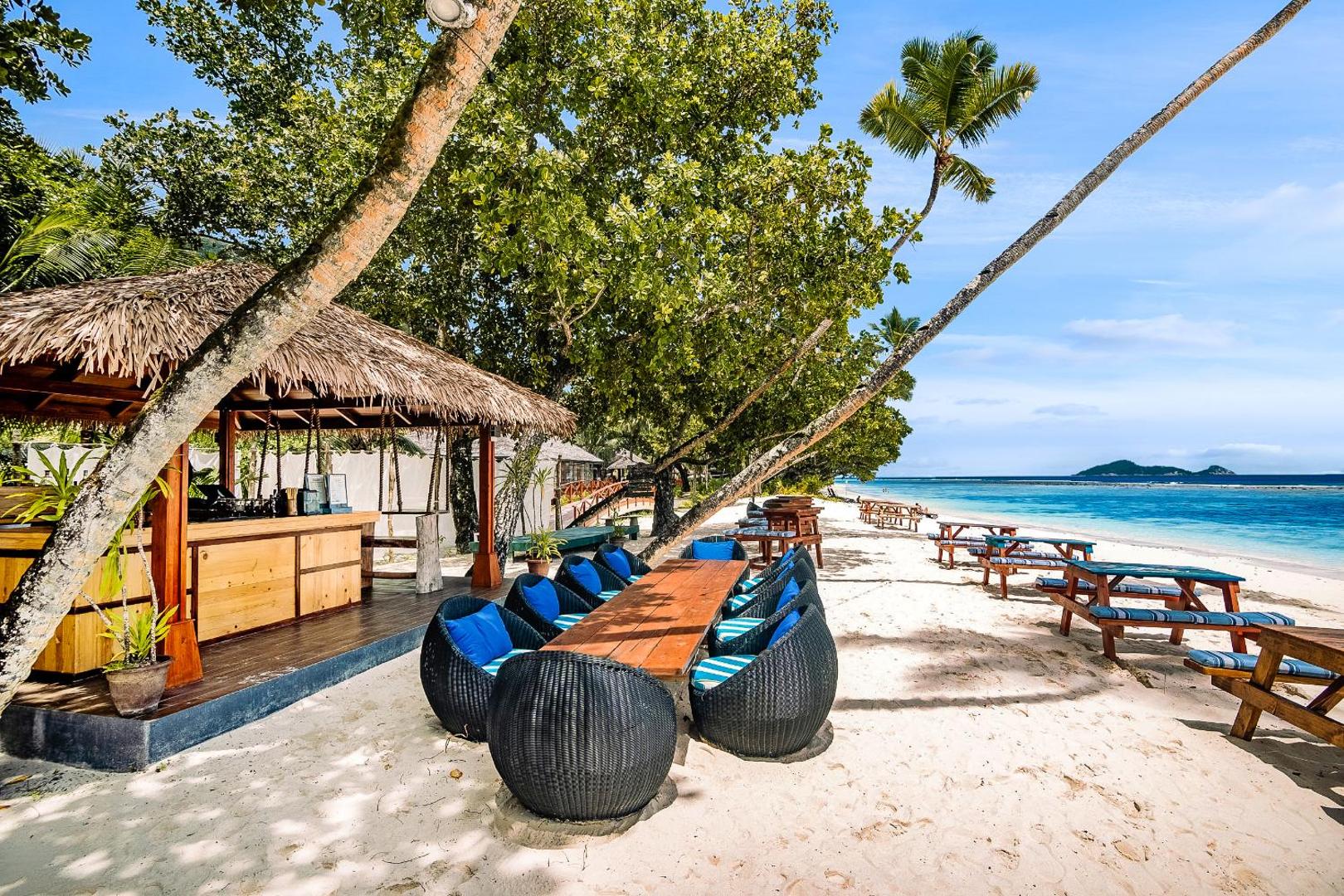 NIVA Labriz Seychelles pludmales bārs ar salmu jumtu un atpūtas zonu smiltīs zem palmām pie zila ūdens
