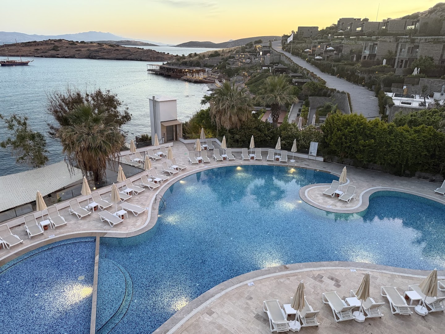 Baska Resort Bodrum baseins vakarā ar izgaismojumu, skatu uz līci un piekrastes apbūvi saulrieta laikā.