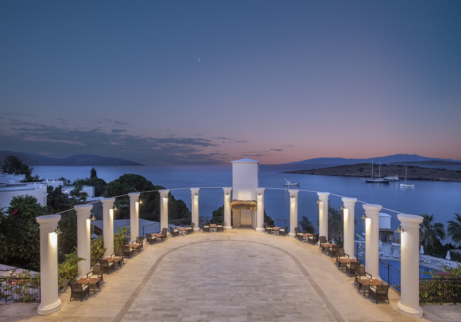 Baska Resort Bodrum terase ar baltām kolonnām krēslas stundā, skats uz Egejas jūras līci un jahtām piekrastē