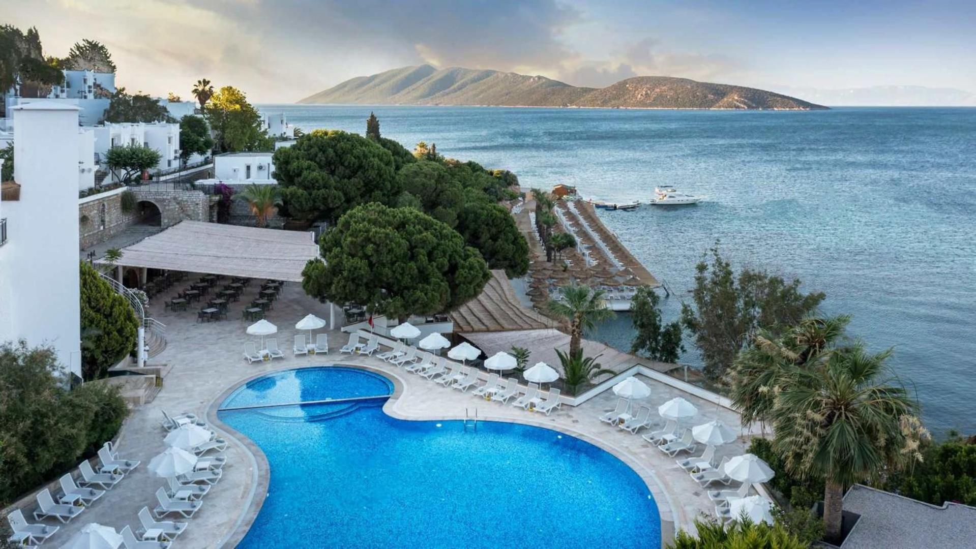 Baska Resort Bodrum viesnīcas panorāmas skats no augšas uz baseinu, terasi un Egejas jūras piekrasti ar kalniem fonā.