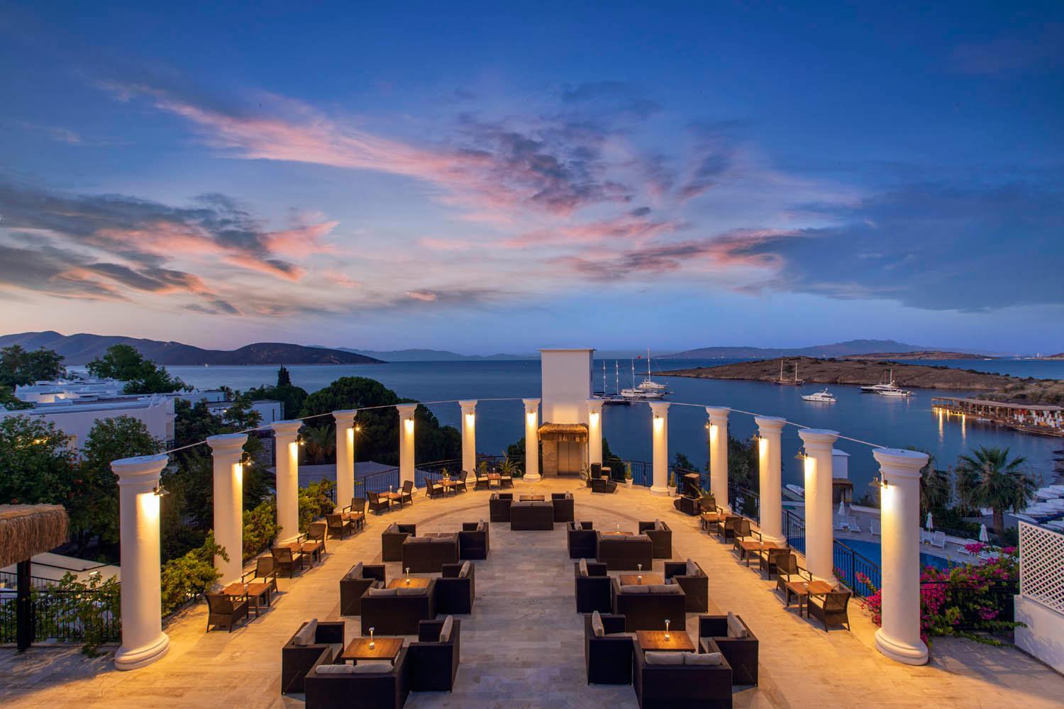 Baska Resort Bodrum āra terase ar atpūtas krēsliem un galdiņiem saulrieta laikā, skats uz jūru un jahtām Bodrumas piekrastē