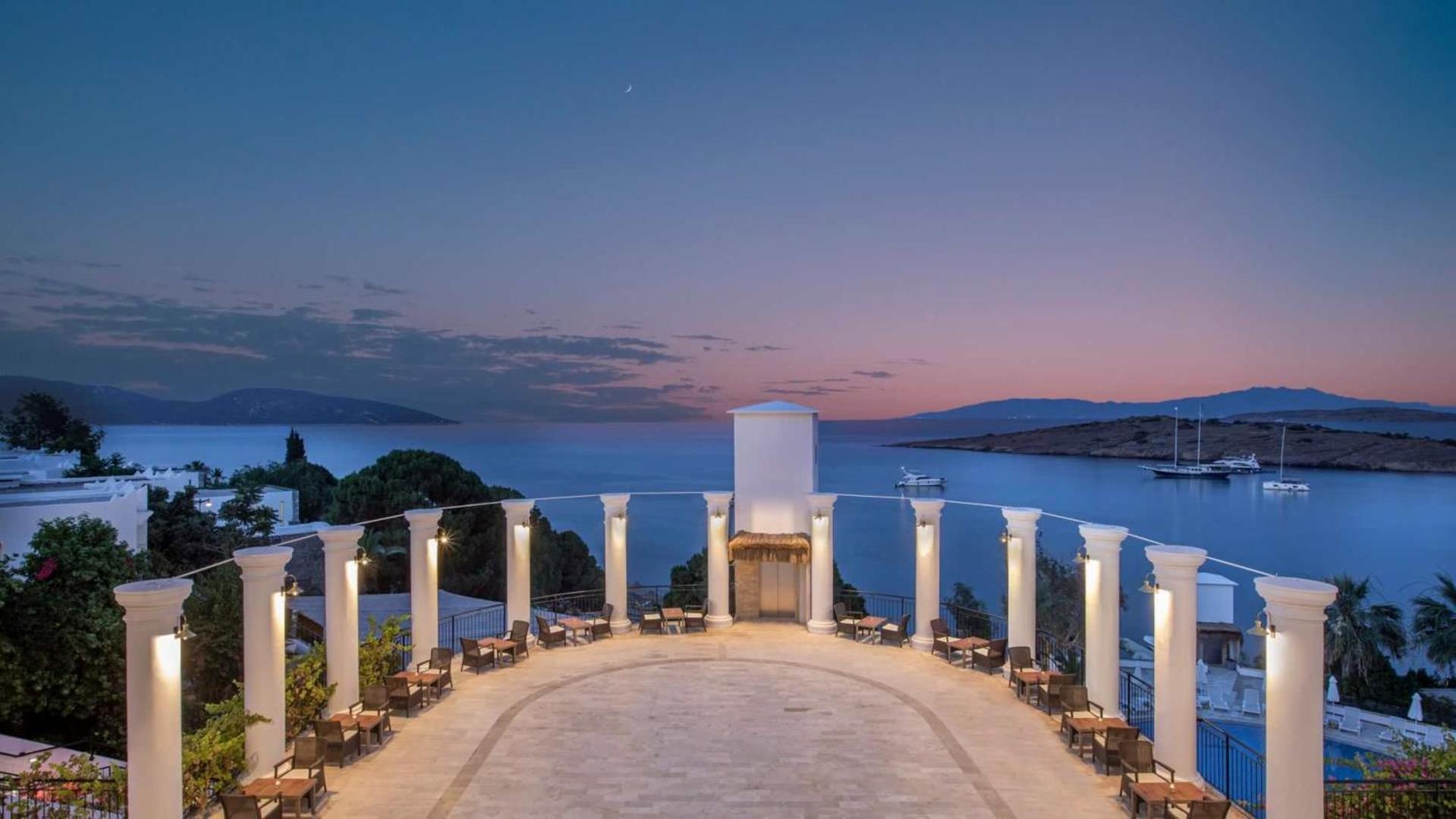 Baska Resort Bodrum terase ar kolonnām un galdiņiem vakara gaismā — skats uz jūru, jahtām un kalniem saulrieta laikā Bodrumā.