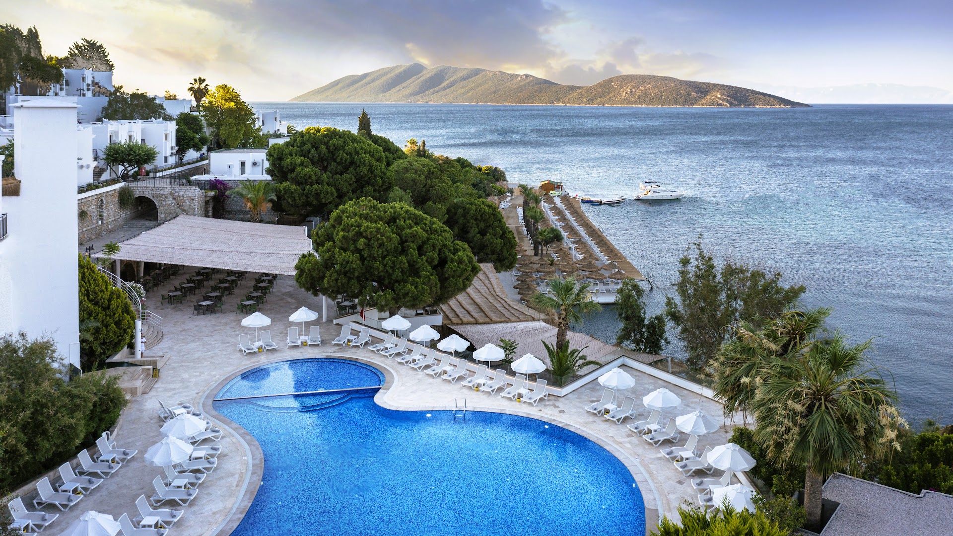 Baska Resort Bodrum teritorijas skats ar baseinu, restorāna zonu un privāto piestātni Egejas jūras krastā saulrietā.