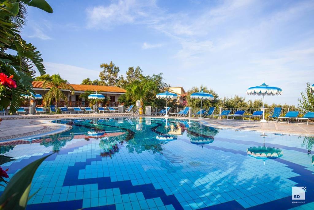 Tonicello Resort & Spa galvenais baseins ar ziliem saulessargiem un viesnīcas ēku fonā zem skaidrām debesīm